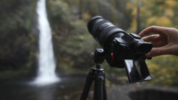Photographer holding a mirrorless camera on a tripod with a waterfall in the background.