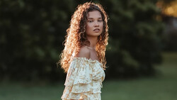 Young woman with long curly copper-toned hair wearing an off-shoulder ruffle blouse in a garden setting.