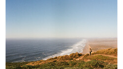 Hiker standing on golden clifftop overlooking a curved coastline with white-capped waves and blue ocean.