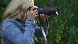 Photographer with blonde hair using a telephoto lens, wearing a denim jacket and sunglasses.