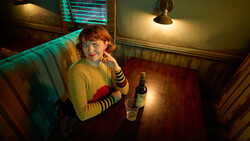 Person with red hair wearing yellow sweater and striped sleeves sits at wooden table with beer bottle under warm and cool mixed lighting.