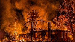 Residential house engulfed in intense flames at night, surrounded by charred trees and heavy smoke.