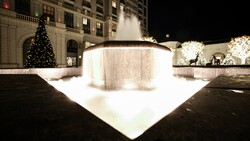 Nighttime courtyard fountain with illuminated basin and cascading water, surrounded by holiday decorations and decorated building facade.