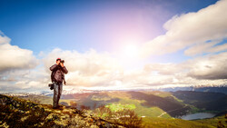 Hiker with backpack stands on a ridge overlooking a vast alpine valley with a lake and snow-capped mountains.