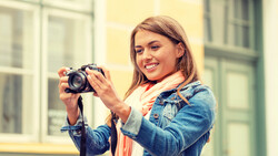 Smiling woman holding a DSLR camera with a telephoto lens, wearing a denim jacket and coral scarf.