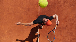 Tennis player executing an overhead serve on a clay court, captured from above with the ball suspended above the net line.