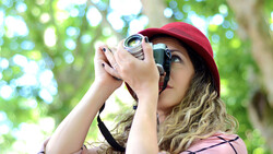 Photographer wearing a burgundy cap and pink shirt looks through a vintage film camera in dappled sunlight.