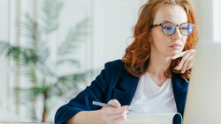 Woman with red hair and glasses working at a laptop while taking notes at a desk.