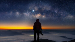 Silhouetted figure standing on a mountain peak facing the Milky Way galaxy at dawn.