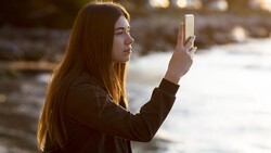 Woman holding smartphone horizontally while standing outdoors near water during golden hour.