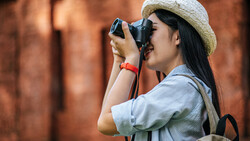 Female photographer wearing a straw hat and light blue shirt holding a camera to her face while photographing against a blurred brick wall background.