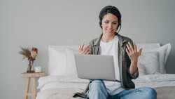 Woman on bed with laptop wearing headset, gesturing while video calling.