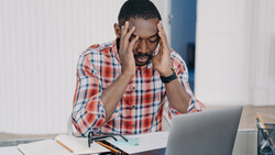 Man in plaid shirt experiencing stress while working at laptop at desk.