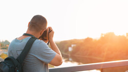 Photographer holding camera to face while standing on bridge at golden hour.