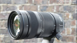 Black telephoto zoom lens mounted on support with brick wall backdrop.