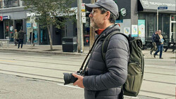 Photographer holding a DSLR camera while wearing a gray cap, black puffer jacket, and olive backpack on an urban street.