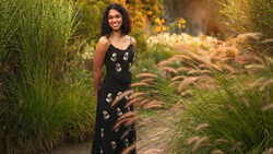 Woman in black floral dress standing among ornamental grasses in golden-hour garden light.