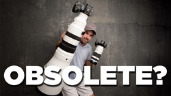 Photographer holding large telephoto lens and camera body overhead against textured gray background.
