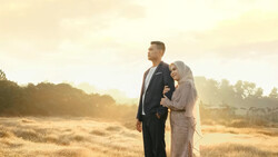 Couple standing together in a sunlit open field at golden hour, looking toward the horizon.
