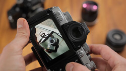Hands holding a mirrorless camera displaying its rear LCD screen with a self-portrait of the photographer holding the same camera.