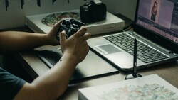Photographer holding a camera controller while editing video on laptop at desk workspace.