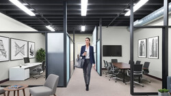 Professional woman in dark blazer walking through modern office corridor with glass partitions and fluorescent lighting.