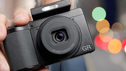 Hand holding a black GR compact camera with a fixed prime lens against a blurred background with colorful bokeh lights.