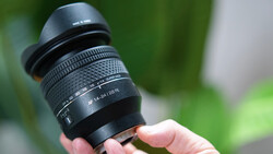 Hand holding a telephoto zoom lens with lens hood attached and visible aperture markings.