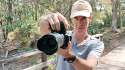 Photographer holding a DSLR with telephoto lens pointed toward camera in a wooded forest setting.