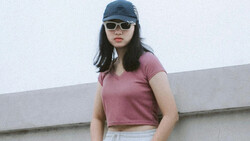 Person wearing pink crop top, white pants, dark cap, and sunglasses against minimalist gray wall backdrop.