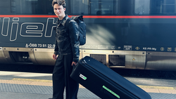 Person in dark formal attire standing beside a black coffin on a train platform next to a passenger train.