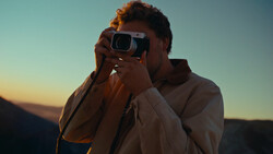 Photographer holding a camera up to his face during golden hour with mountains in the background.