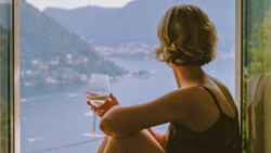Woman holding a wine glass overlooking a mountainous lake landscape from an interior vantage point.