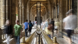 Blurred figures walking through a classical colonnade with arched ceilings and ornate columns.