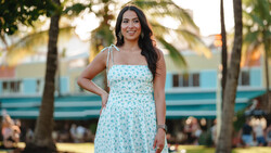 Woman in light blue floral dress standing in park with palm trees and colorful building behind her.