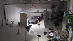 Professional car photography studio setup with luxury sedan, overhead softbox lighting, and crew monitoring shoot.