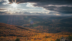 Autumn forest landscape with layered mountains beneath dramatic sky with sun rays.
