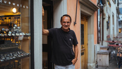 Man in black shirt leaning against doorframe of watch shop at dusk.