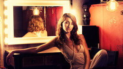 Woman seated at vanity mirror with warm bulb lighting, gazing toward camera in studio setting.