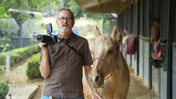 Photographer with vintage film camera beside a brown horse in a stable corridor.