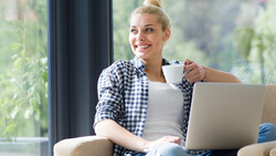 Woman with blonde hair in bun wearing blue gingham shirt, holding white mug while looking at laptop by window.