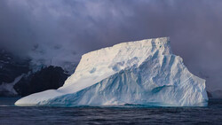 Large tabular iceberg with steep blue-tinted faces floating in dark Antarctic waters.