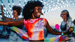 Woman wearing colorful gradient tank top and headphones dances outdoors at twilight under string lights.