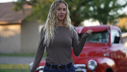 Woman with long blonde wavy hair wearing a dark long-sleeve shirt and jeans, standing outdoors with a vintage red truck blurred in the background.
