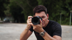 Male photographer holding a DSLR camera with telephoto lens, looking through the viewfinder.