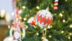 Red and white patterned ornament hanging from evergreen branch with warm bokeh lights in background.