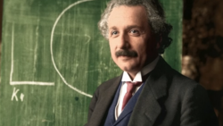 Man in formal attire with mustache standing before green chalkboard with geometric diagram.