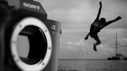Sony α7 camera body in foreground with silhouetted person diving into water near sailboat in background, black and white.