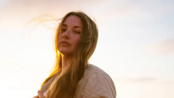 Woman with long blonde hair backlit by warm golden sunset light against a soft, overcast sky.