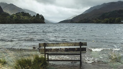 Weathered wooden bench partially submerged in calm lake water, facing mountains under overcast sky.